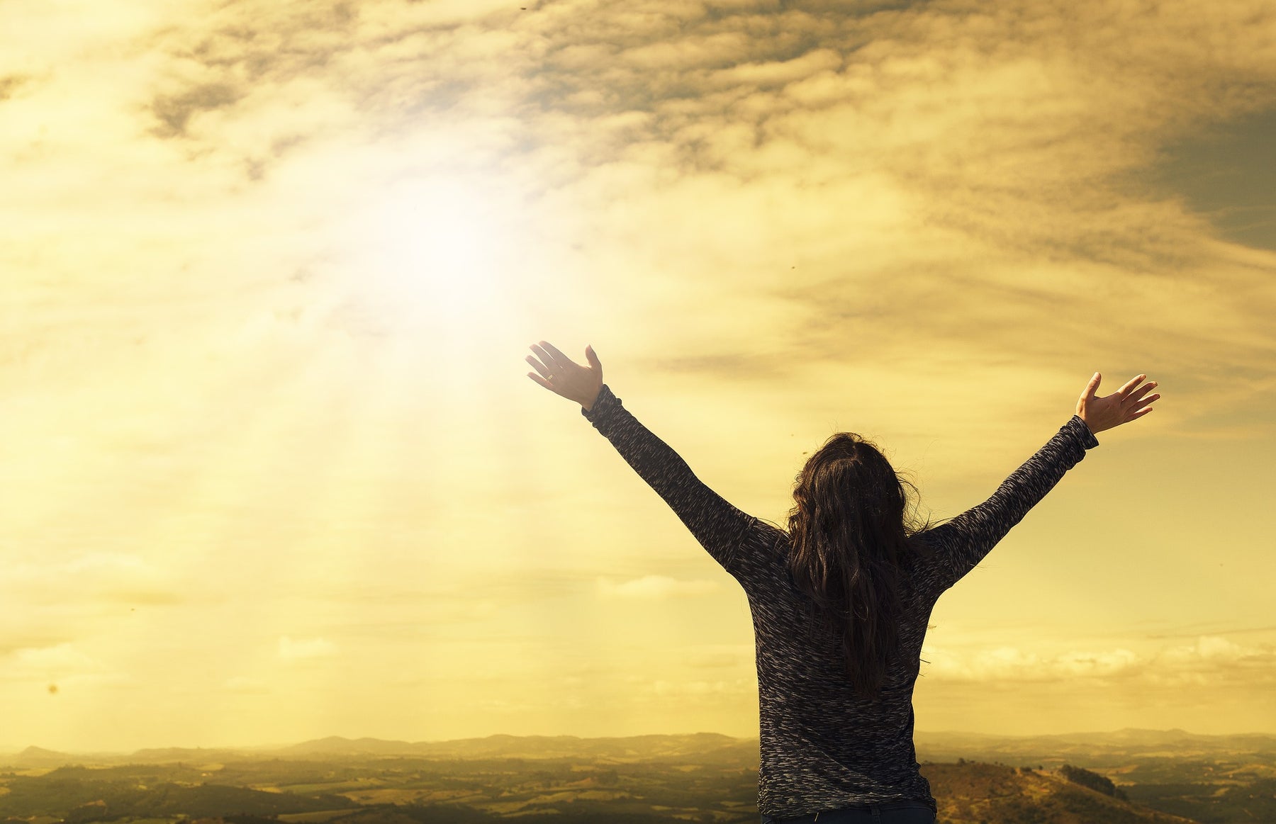 Woman with rised hand looking at the sun coming out of clouds over a field